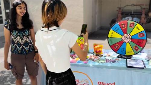 Two smiling young women at outdoor booth with colorful prize wheel and snacks.